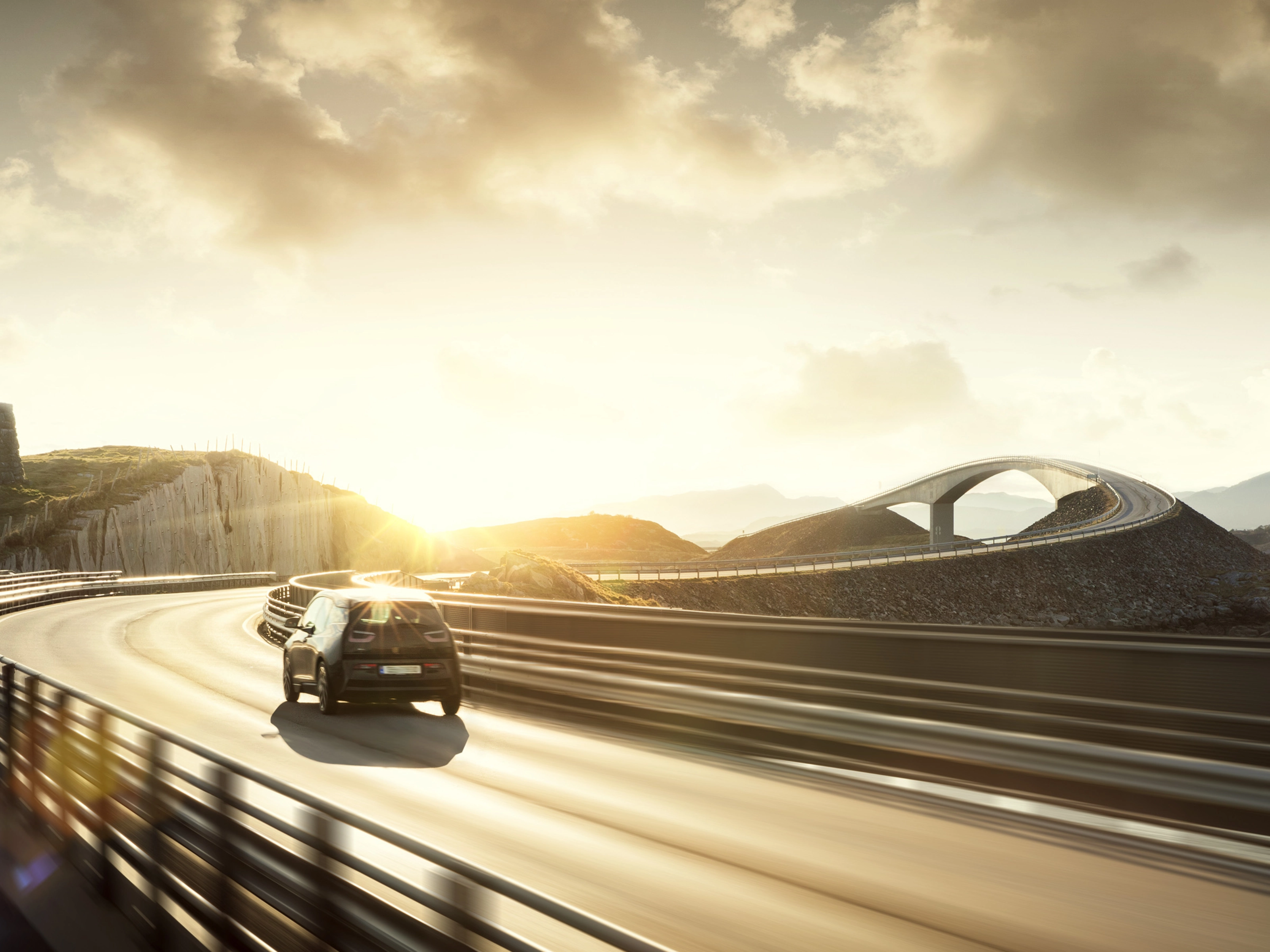 Ein Auto fährt bei Sonnenuntergang über eine kurvige Brücke in einer Küstenlandschaft.