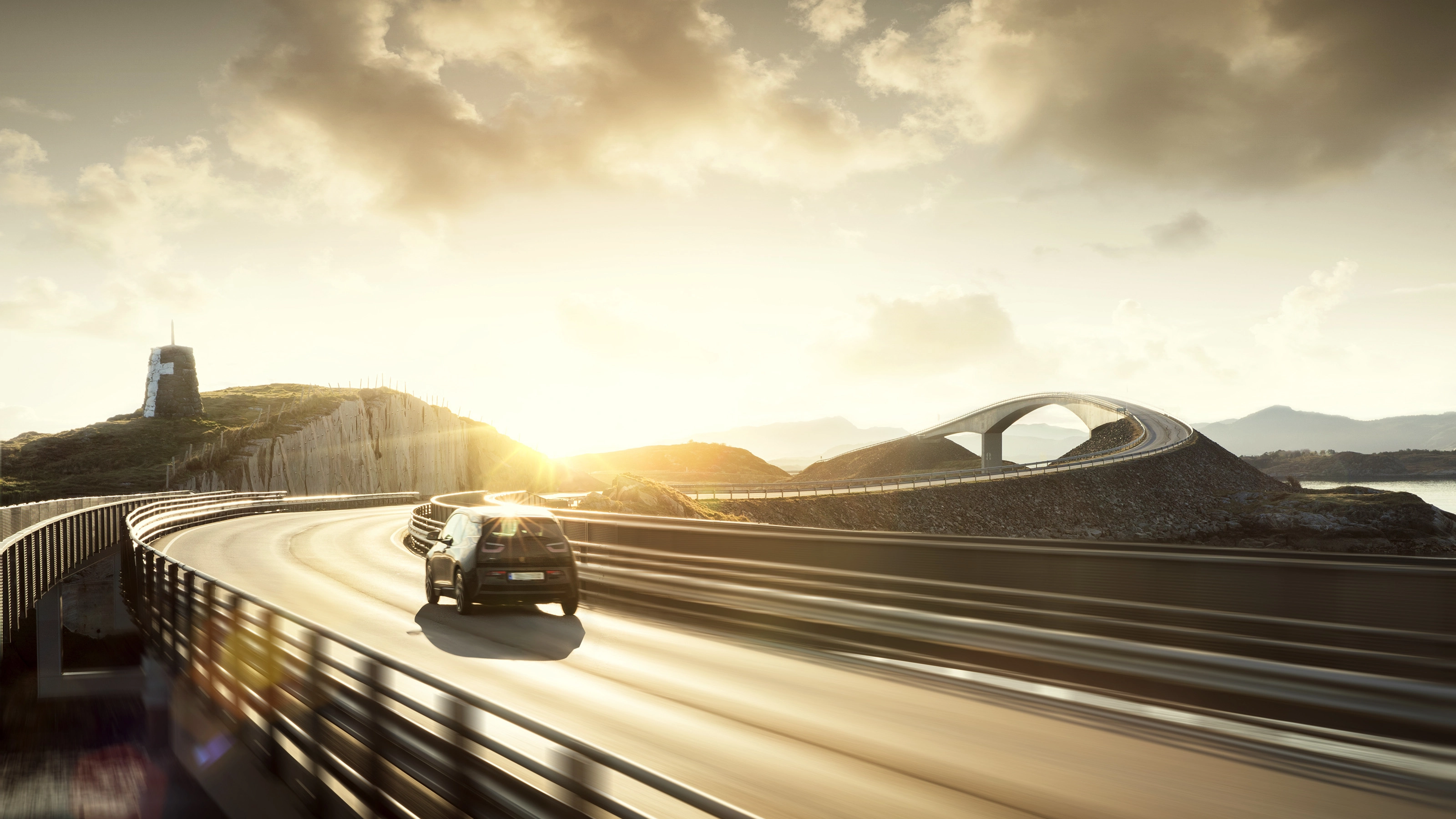 Ein Auto fährt bei Sonnenuntergang über eine kurvige Brücke in einer Küstenlandschaft.